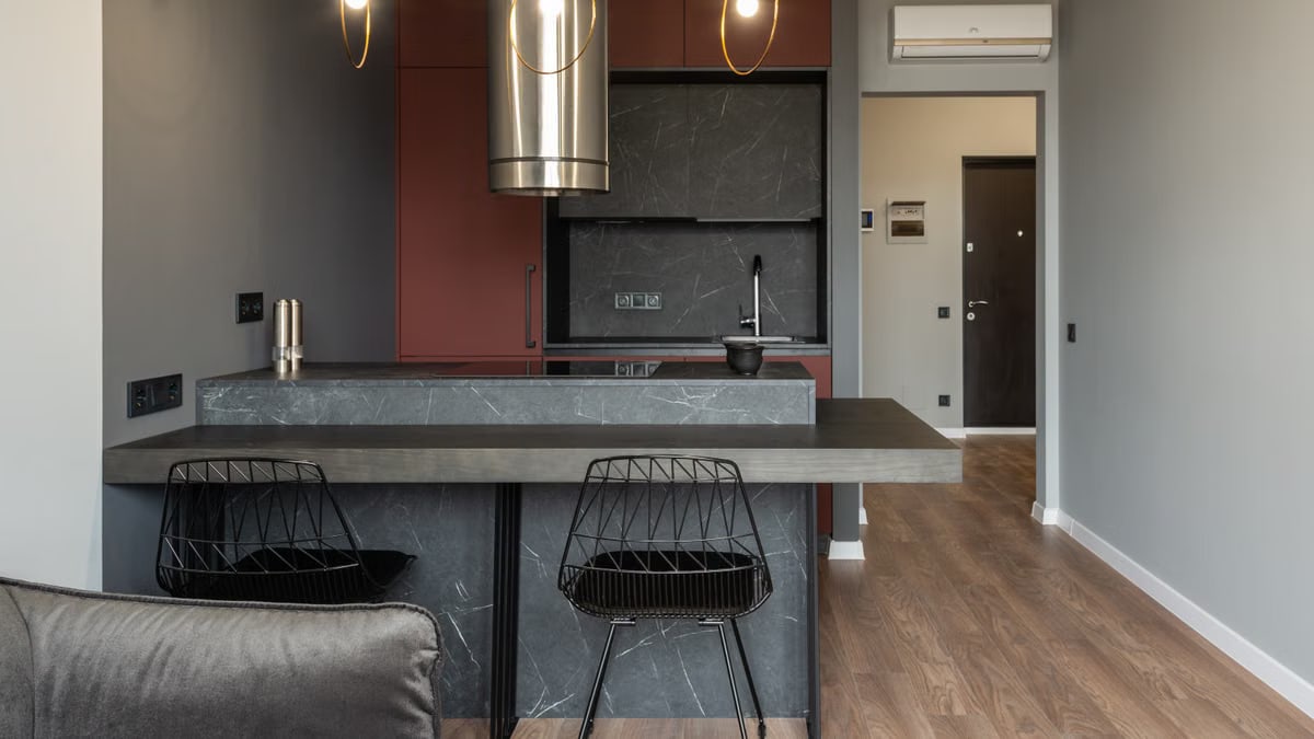 Dining zone furnished with counter and chairs in modern apartment with island style kitchen equipped with range hood and illuminated by hanging lamps